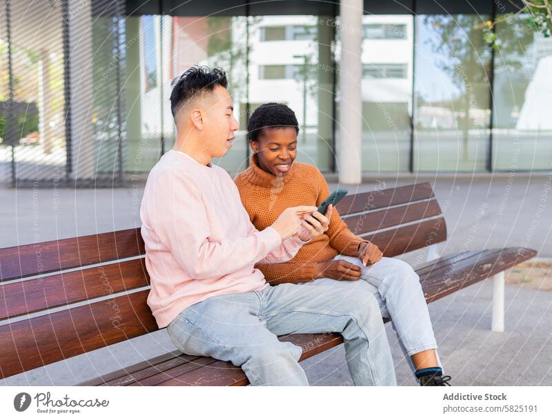 Two friends sharing a moment with a smartphone on a bench friendship outdoor asian man african american technology connection smiling focused social media