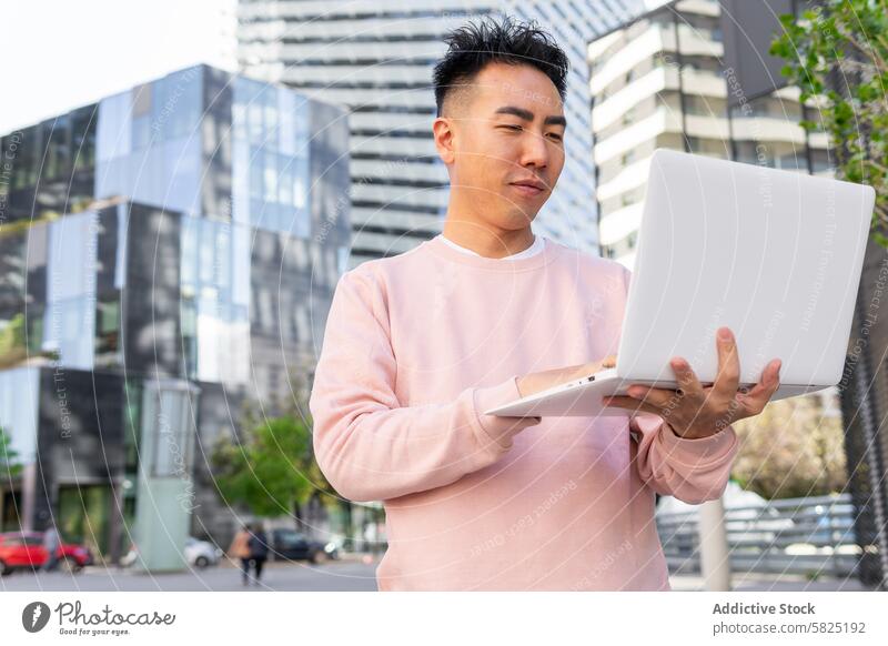 Young Asian man using a laptop outdoors in the city young asian modern buildings casual clothing pink sweater standing technology gadget using device day sunny