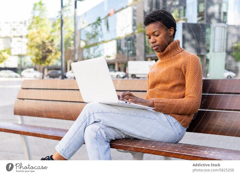 African American student working on laptop outdoors woman african american bench focused technology sitting engrossed task urban casual sweater jeans park