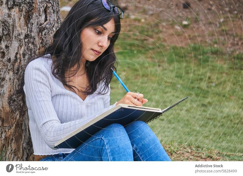 Serene Moment of a Woman Writing in the Park woman writing park tree notebook creativity lush serene engrossed concentration peaceful leisure activity outdoor