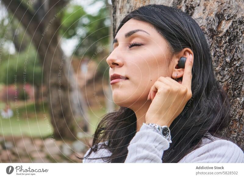 Serene woman enjoying a peaceful moment in the park relaxing tree serene wireless earbud tranquility lush eyes closed nature outdoor leisure relaxation calm