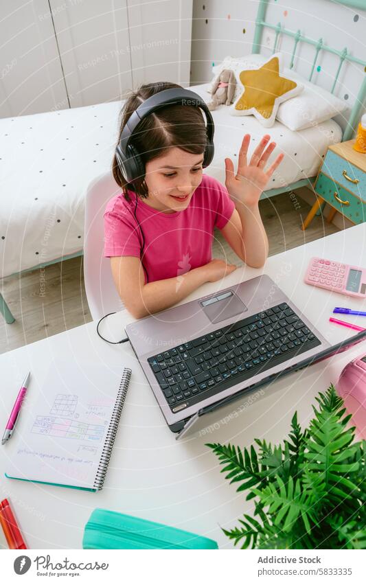 Young girl engaging in online learning from home child education virtual class bedroom home school laptop headphone wave cheerful colorful study youth