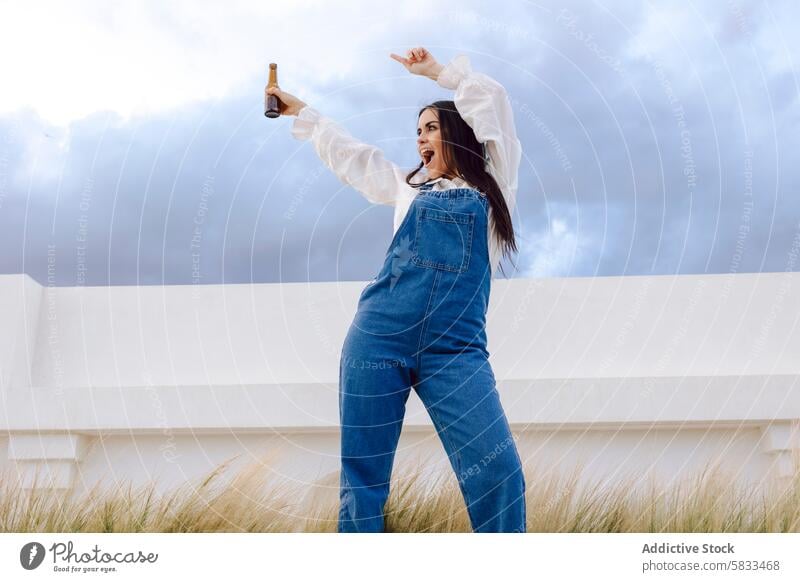 Celebratory woman with beer at the port joy celebration bottle denim overalls sky clouds festive pose holding alcohol leisure dock waterfront maritime fun young