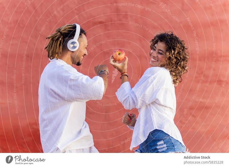 Two joyful people exchanging an apple outdoors man woman playful smile casual white top red wall fruit exchange sharing friendship enjoyment happiness headphone