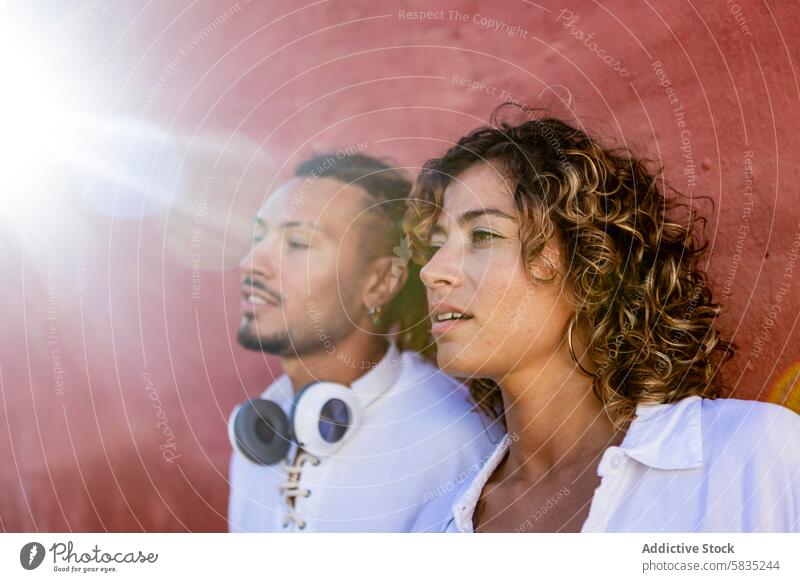 Young couple in casual wear gazing into distance young stylish thoughtful looking woman headphones curly hair modern style vibrant red wall daytime outdoor