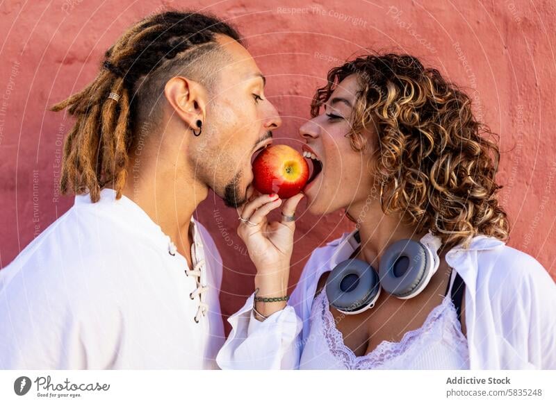 Young couple sharing an apple against a colorful wall young playful bite man woman joy red wall casual outfit white shirt dreadlocks curly hair joyfulness