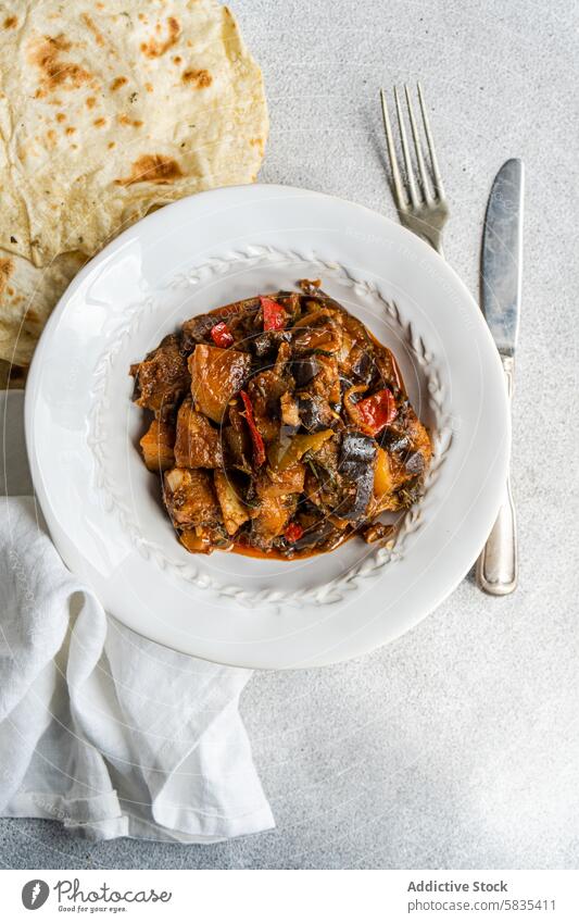 Homemade vegetable stew and flat bread on a white plate eggplant tomato potato bell pepper onion garlic basil homemade nutritious meal fork knife napkin hearty