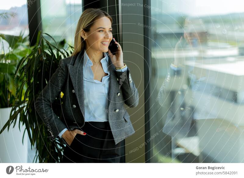 Smiling businesswoman talking on mobile phone by office window in daylight conversation grey blazer confident smiling reflection professional female attire