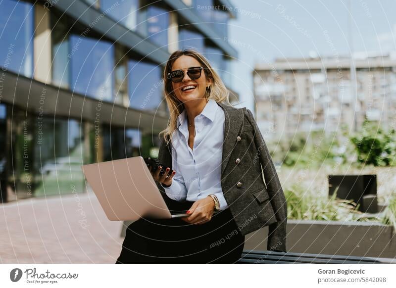 Professional businesswoman working on laptop outside modern office building in daylight architecture bench blazer career city communication confident