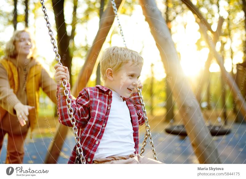 A mother swings her son on a swing in an autumn city park. A beautiful woman entertains her adorable baby on the playground. together yellow leaves having fun