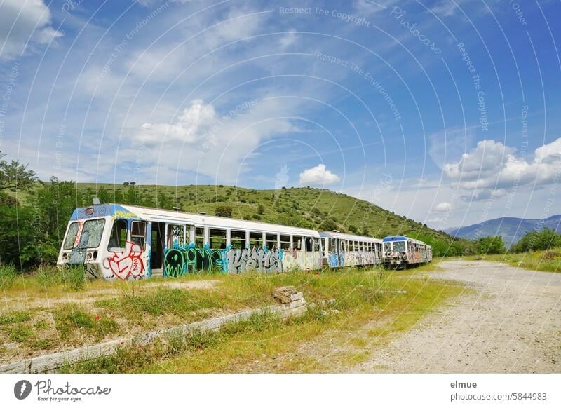 Decommissioned, destroyed railcars with graffiti in a green landscape with fair weather clouds in the blue sky / terminus station Railcar Motorized trolley