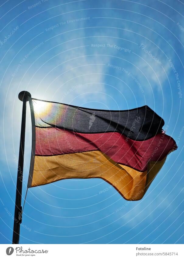 The flag of Germany flies in front of a blue sky with light clouds on the flagpole. The sun is shining through the fabric. Flag German flag German Flag Ensign