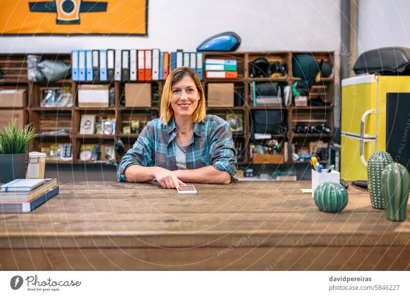 Portrait of smiling blonde young female employee looking at camera behind of industrial shop counter portrait happy woman freckles wear plaid shirt working