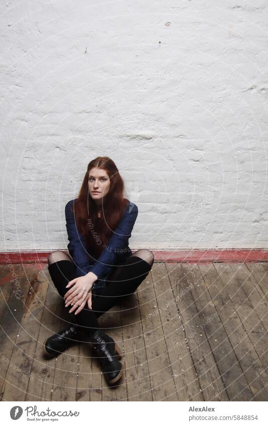 Very tall, young woman sitting on a plank floor in front of a white wall with a red baseboard Woman tall woman very large pretty Long-haired Brunette