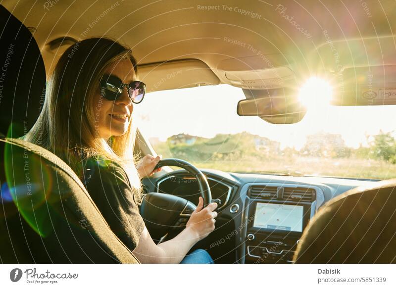 Woman wearing sunglasses is driving car with sunlight through windshield woman driver sunny steering wheel vehicle smile daylight road trip rural portrait