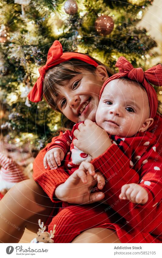 Sibling love during festive Christmas celebration christmas siblings red tree decorated embracing joy camera girl baby smile bow outfit headband matching family
