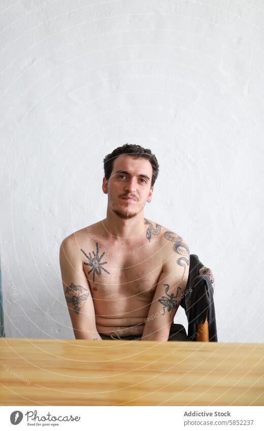 Tattooed young man posing with a contemplative expression portrait tattoo shirtless wood table white background thoughtful ink body art skin pose sitting