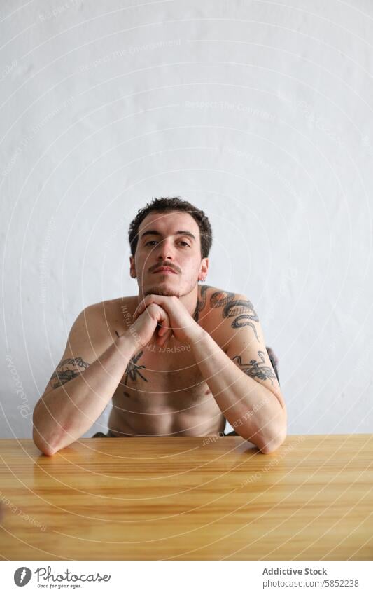 Shirtless tattooed young man sitting at a wooden table male shirtless white background thoughtful gaze intricate torso arm skin art casual home indoor