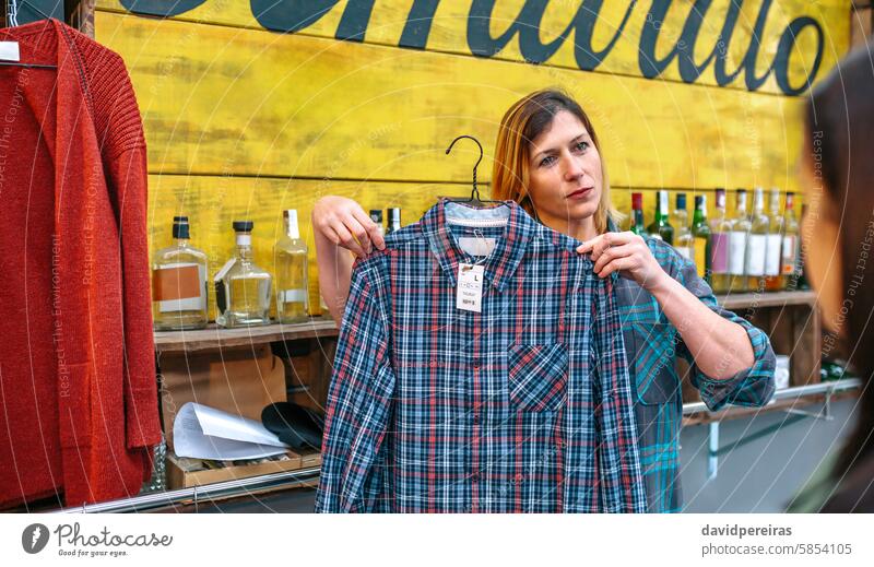 Friendly woman shop assistant with freckles showing plaid shirt to customer in store. client consumer female looking clothes second hand local market