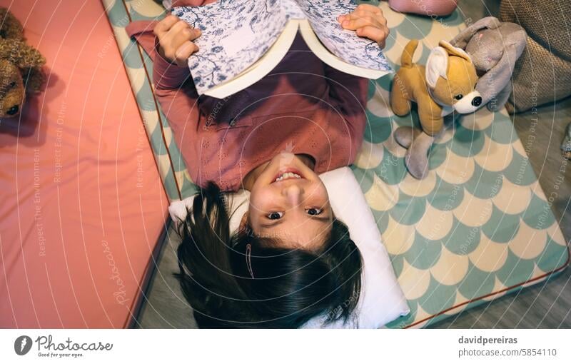 Top view of smiling little girl looking at camera while holding book and resting over sleeping mats happy smile reading lying mattress living room stuffed dog