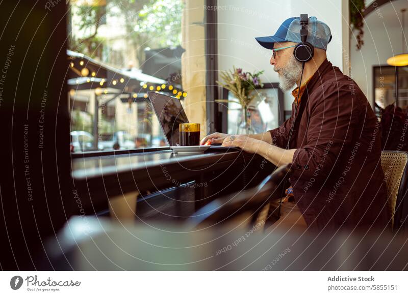 Modern senior man working remotely in a cafe setting modern remote work lifestyle elderly laptop headphones technology baseball cap stylish cozy indoor
