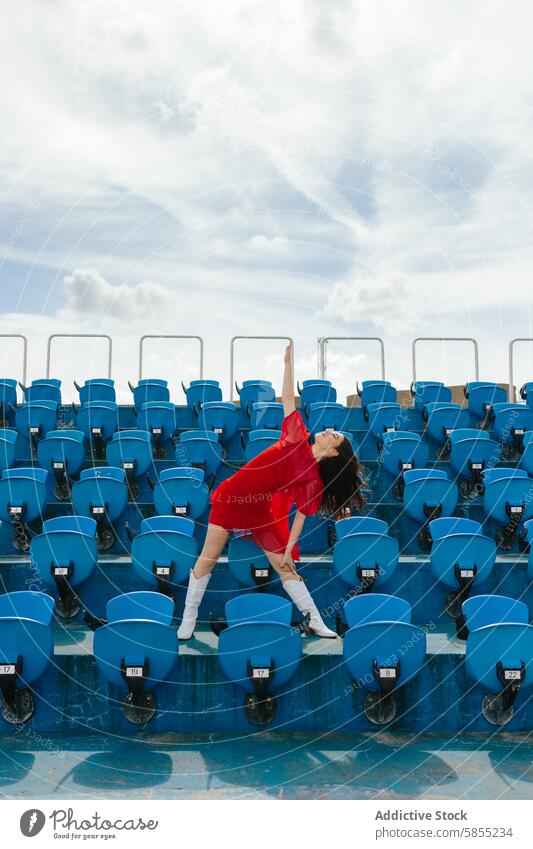 Young woman dancing joyfully in a stadium dance seat red dress blue sky cloud energy youthful movement outdoor celebration fashion style happy vibrant leisure