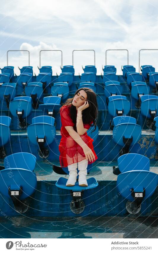 Young woman in red dress sitting alone in stadium seats blue seat thoughtful day outdoor solitude young fashion style boots white boots casual leisure summer