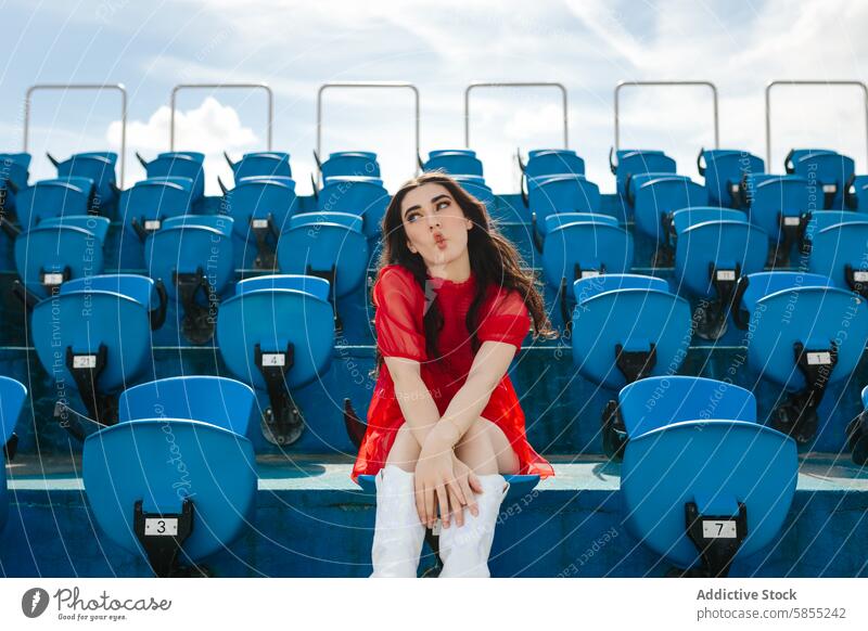 Young woman alone in a stadium sending a kiss seat red blue playful outdoor sky day summer young casual fashion style urban seating empty event solitude leisure