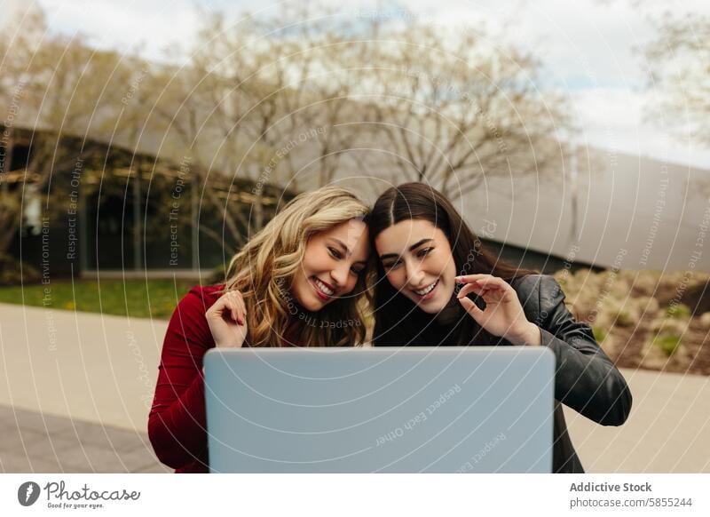 Two friends sharing a joyful moment with a laptop outdoors woman park technology smile laugh looking screen young adult female friendship happy happiness