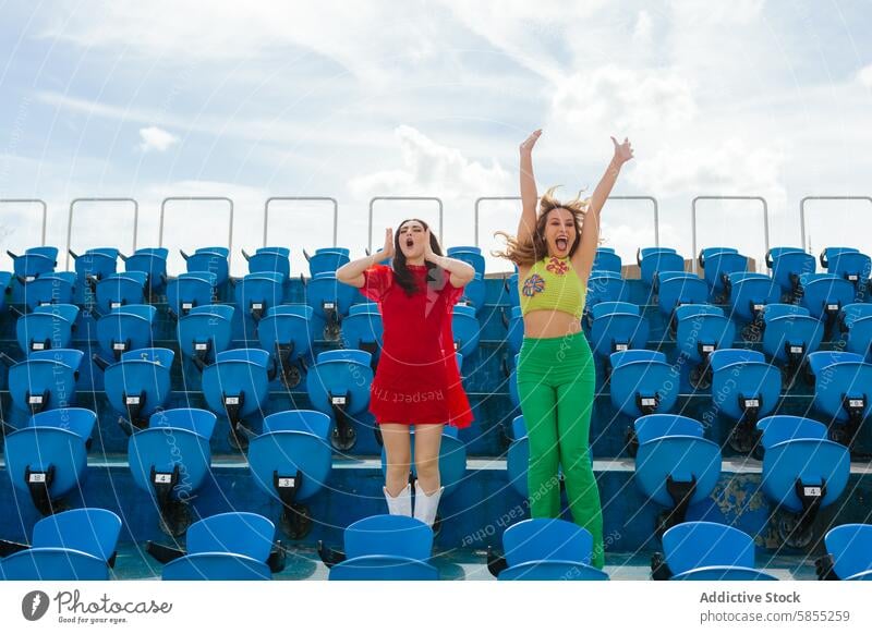 Young women cheering enthusiastically in a stadium woman jump energetic empty seat blue sky vibrant outfit female friend joy excitement celebration leisure