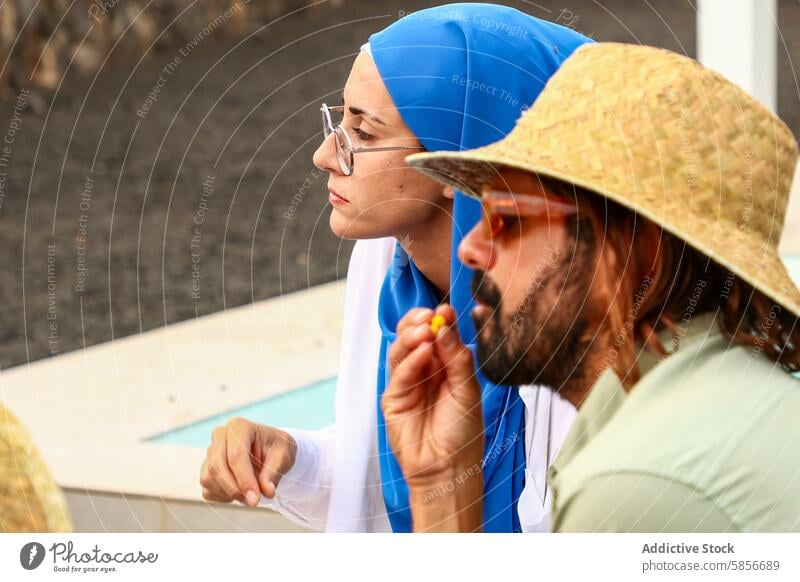 Woman in blue headscarf looks at camera, man grazes beside her woman muslim straw hat sunglasses snack outdoor thoughtful contemplative direct gaze looking away