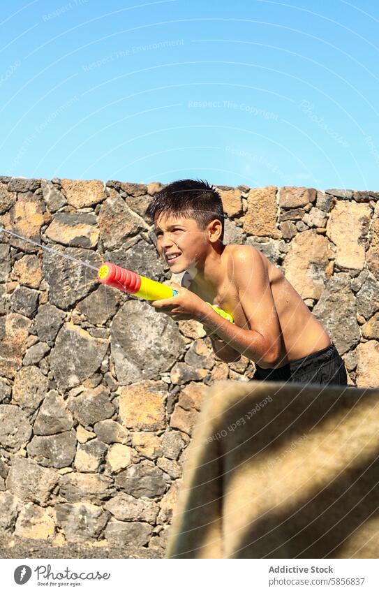 Boy playing with water gun by stone wall boy smile looking away outdoor sunny summer fun leisure activity childhood squirt wet joy playful sunshine warm happy