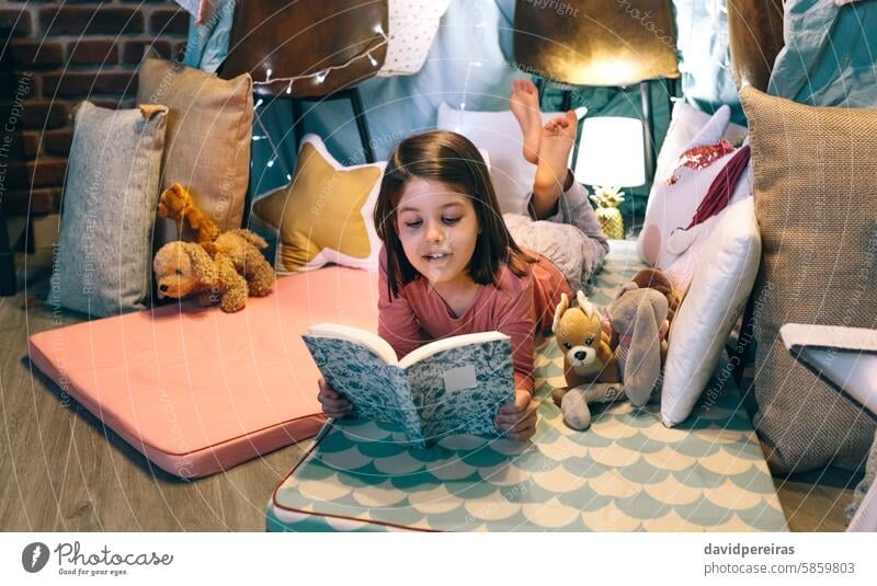 Happy little girl reading and relaxing lying over sleeping mat in a cozy tent at home happy book mattress serene child resting smile holding cushions surrounded
