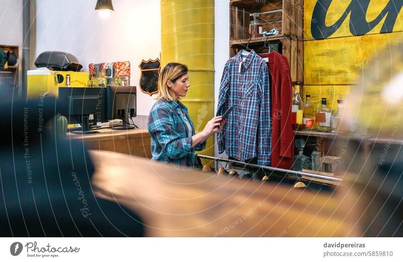 Blonde young woman looking digital tablet while checking stock on second hand clothing store holding clothes female working shopping industrial style