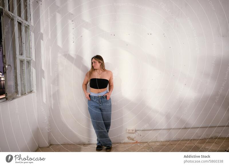 Young woman posing in an industrial studio setting casual leaning wall rustic fashion style youth female denim jeans tube top urban modern casual wear lifestyle