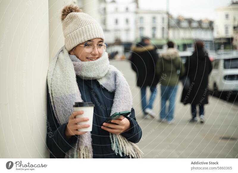 Smiling woman with coffee and smartphone on London street london fashion accessory winter smile technology city life urban glasses scarf hat drink beverage