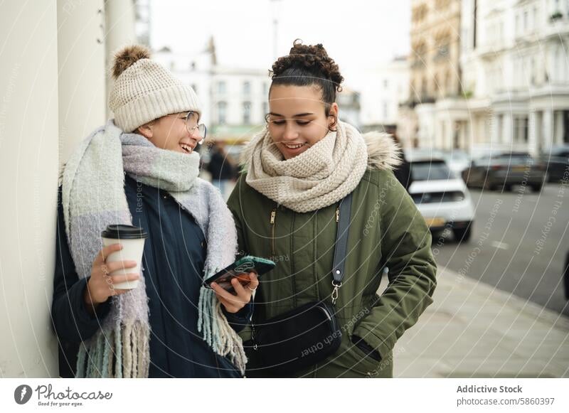 Two women enjoying a chat with smartphones in London london street coffee smile urban connectivity young winter scarf jacket city casual friendship technology