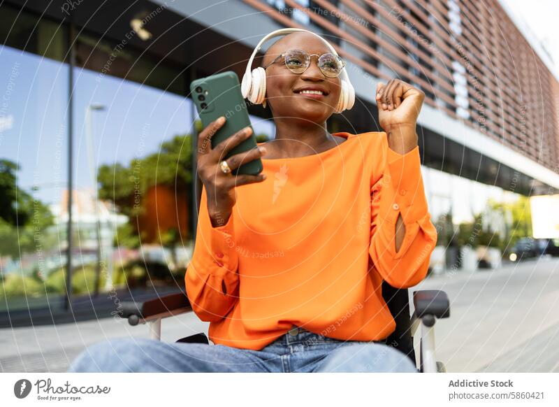 Joyful businesswoman in wheelchair using smartphone outdoors urban sunny confident happy music headphone orange blouse building modern technology accessibility