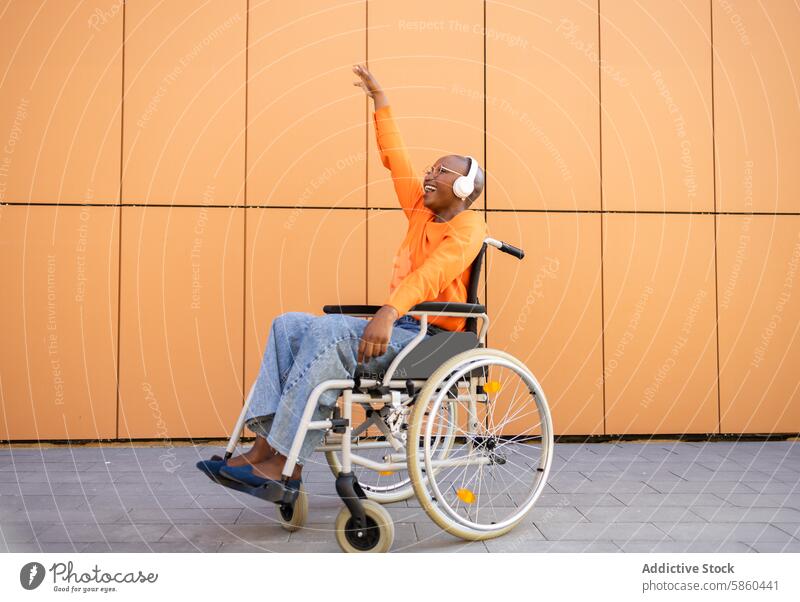 Joyful woman in wheelchair reaching upwards outdoors joy cheerful black woman orange top jeans headphones hand muted orange background accessibility mobility