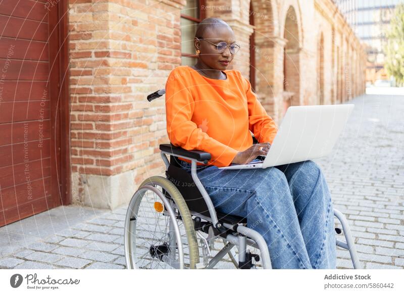 African American woman in wheelchair working on laptop outdoors business african american black ethnic accessibility inclusion street brick orange jeans glasses