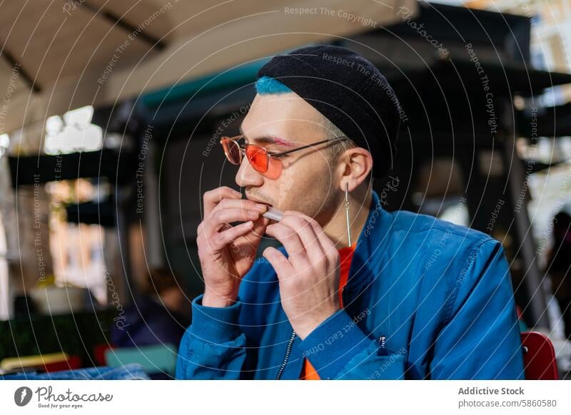 Young man eating hamburger in street cafe with stylish look young blue jacket teal hair black beanie orange glasses outdoor dining casual fashion trendy meal