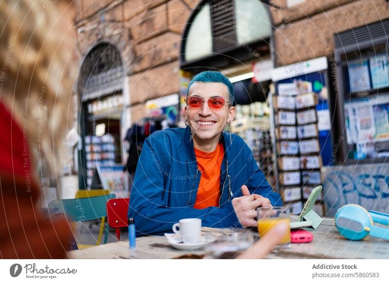 Young man enjoying coffee at a street cafe in the city young outdoor smile digital device book urban table blue hair red sunglasses cheerful daylight
