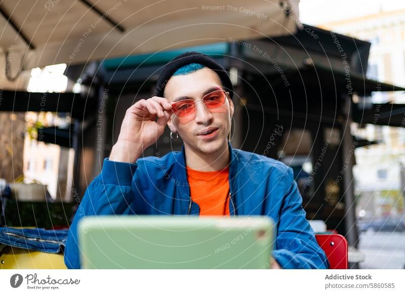 Stylish young man adjusting sunglasses at cafe stylish outdoor laptop fashion urban casual sitting touch rose-tinted blue jacket orange shirt hat daylight city