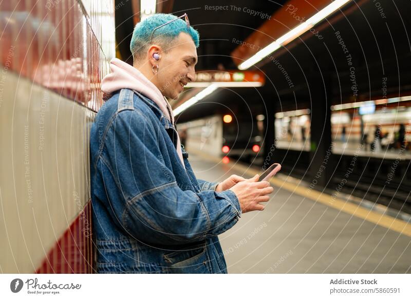 Young man with blue hair using smartphone in subway station young waiting train denim jacket in-ear headphone mobile technology urban casual modern lifestyle