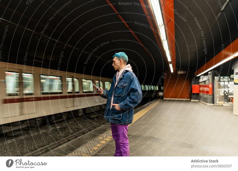 Young man with blue hair waits for train while using phone subway platform transportation urban commute waiting jacket standing wireless communication