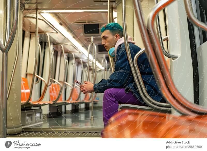 Young man with blue hair enjoying music on subway ride young headphone smartphone cheerful denim jacket purple trouser public transport seating absorbed joyful