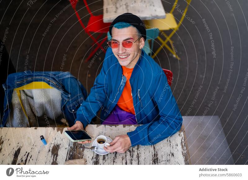 Young man enjoying coffee outdoors at colorful table young cafe smartphone blue hair red sunglasses smiling street chair urban style casual jacket denim sitting