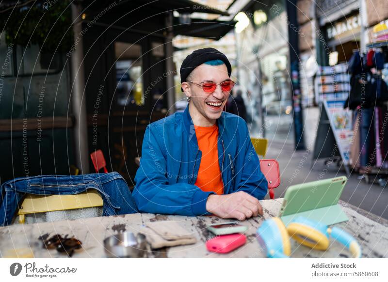 Stylish young man laughing at a cafe with smartphone and headphones outdoor table beanie sunglasses red blue jacket joy happiness lifestyle urban street casual