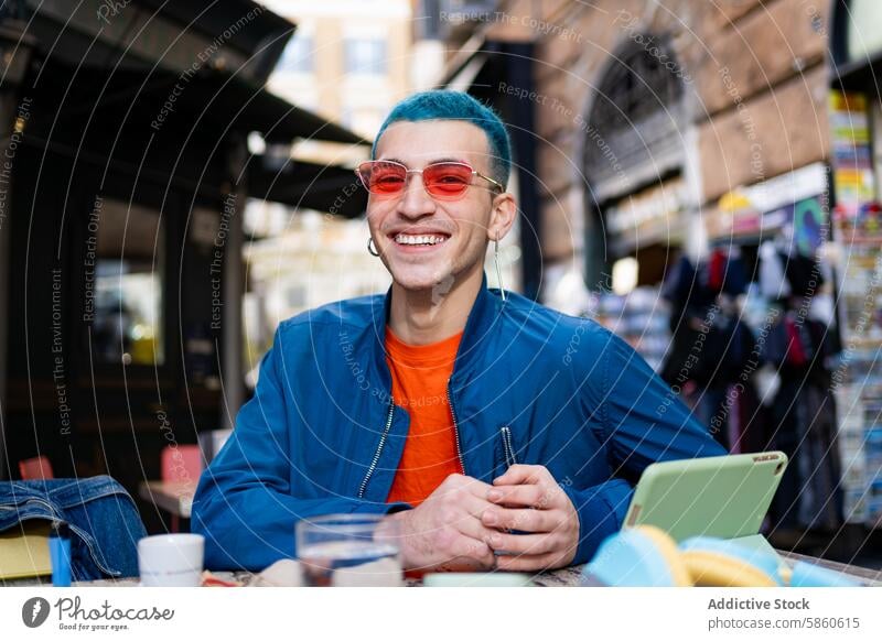Smiling young man with blue hair enjoying coffee outdoors smile red sunglasses cafe smartphone table happiness fashion style urban street casual adult male