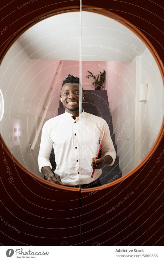 Smiling businessman seen through a circular mirror on stairs smile happy reflection document indoor office corporate african white shirt professional standing